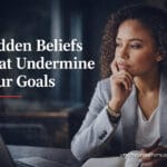 Mixed race professional woman reflecting at desk with Hidden Beliefs That Undermine Your Goals blog title overlay